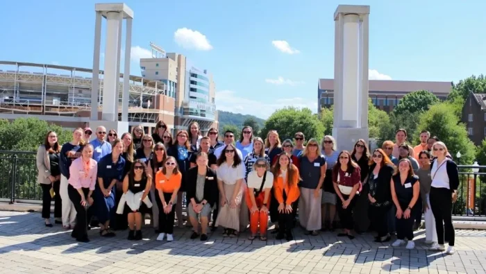 Large group of BisNet 2024 participants dressed in casual clothing - some in UT orange - pose on UT's pedestrian bridge with Neyland Stadium in the background|Large group of BisNet 2024 participants dressed in casual clothing - some in UT orange - pose on UT's pedestrian bridge with Neyland Stadium in the background||