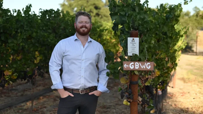 Charles Dick stands smiling in a California vineyard. He has red hair and a red beard and mustache. He is wearing a light