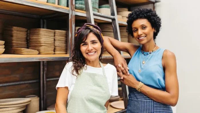 Two female business owners stand among shelves of pottery.